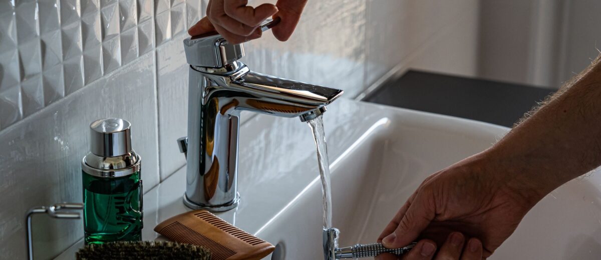 A man's hands adjust the faucet in a bathroom, surrounded by grooming tools like a comb and brush.