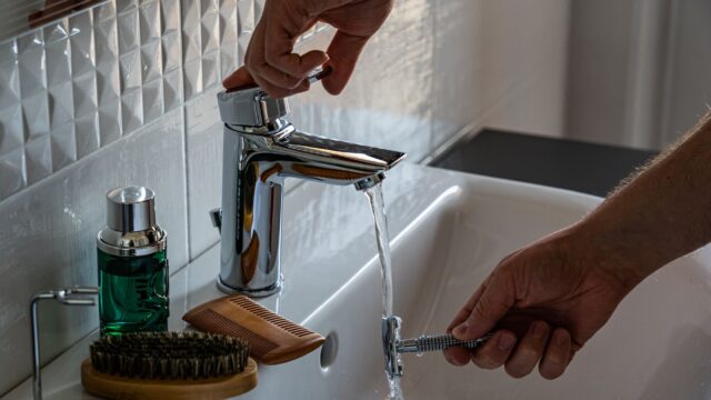 A man's hands adjust the faucet in a bathroom, surrounded by grooming tools like a comb and brush.